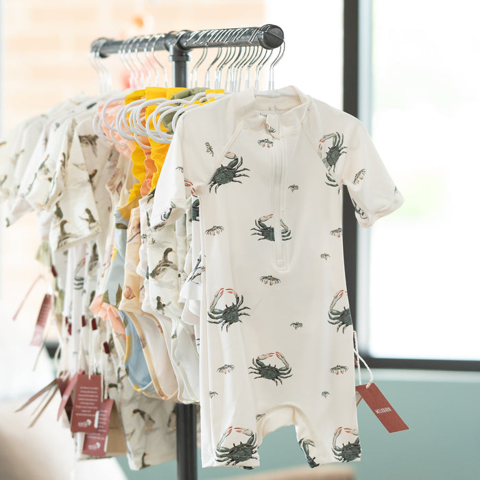 Baby rashguard swimsuitss with crab pattern on a clothing rack against a window background