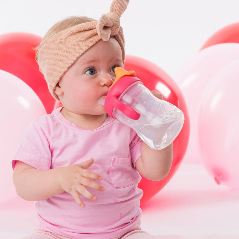 Baby in pink outfit drinking from a sippy cup with balloons in the background