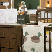 A nursery furniture display featuring a dark brown wooden dresser and crib with a cream knit blanket draped over the crib, printed with a green and brown duck pattern. A plush duck toy sits on a black branded display box on the dresser.