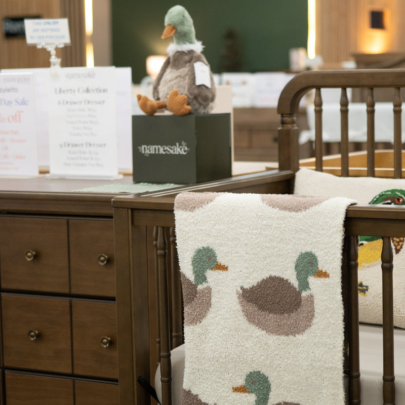 A nursery furniture display featuring a dark brown wooden dresser and crib with a cream knit blanket draped over the crib, printed with a green and brown duck pattern. A plush duck toy sits on a black branded display box on the dresser.