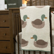 A cream-colored plush blanket with a repeating duck pattern draped over a wooden crib in a nursery setting. A plush duck toy sits on a dark green box labeled 'namesake' on the crib's side.