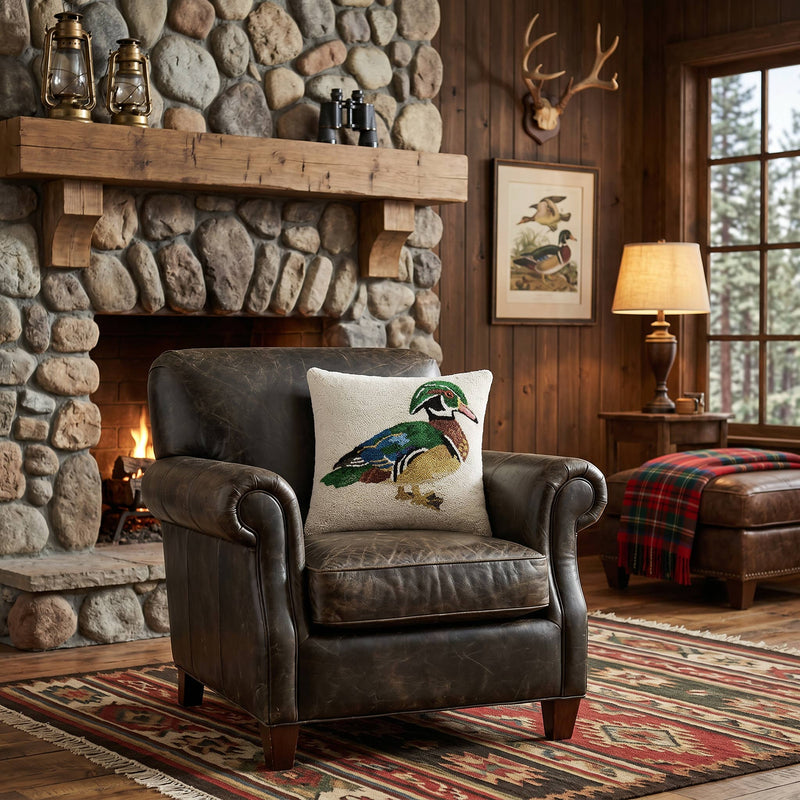 A rustic brown leather armchair in a cabin setting, featuring a cream throw pillow with a colorful wood duck illustration. The chair is positioned in front of a stone fireplace with a wood mantel, a lit fire, and a patterned rug.