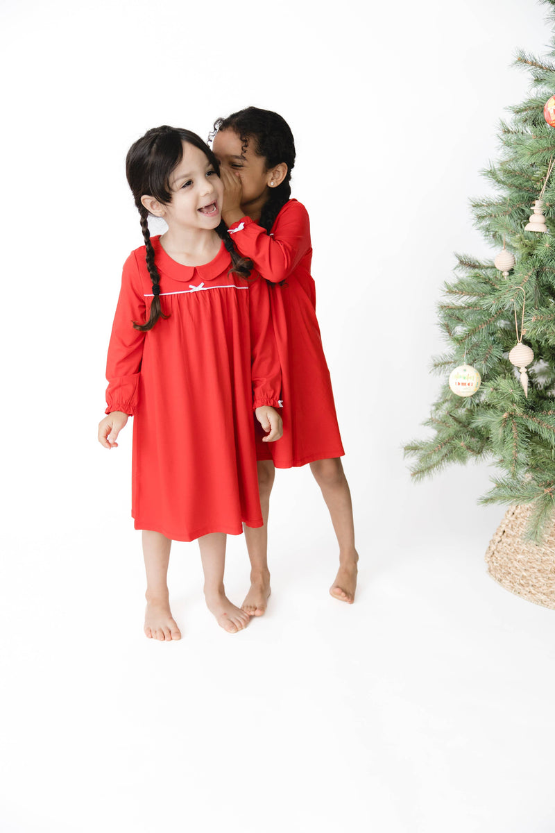 Two young girls wearing matching red long-sleeve dress sleepwear stand barefoot on a white studio background, with one girl whispering to the other. A decorated Christmas tree is visible on the right side of the frame.