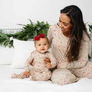 Mom and baby wearing matching Lou Lou & Co Holly print pajamas by a Christmas garland