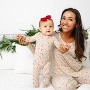 A smiling woman and a baby wearing matching cream-colored footed pajamas with a small red floral print, sitting on a white bed with white pillows and green plants in the background.