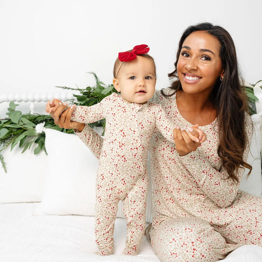A smiling woman and a baby wearing matching cream-colored footed pajamas with a small red floral print, sitting on a white bed with white pillows and green plants in the background.