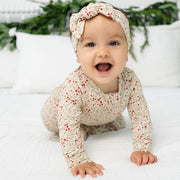 A baby crawling on a white quilted bed wearing a cream long-sleeve jumpsuit and matching headband with a red and green berry pattern, with white pillows and green foliage in the background.