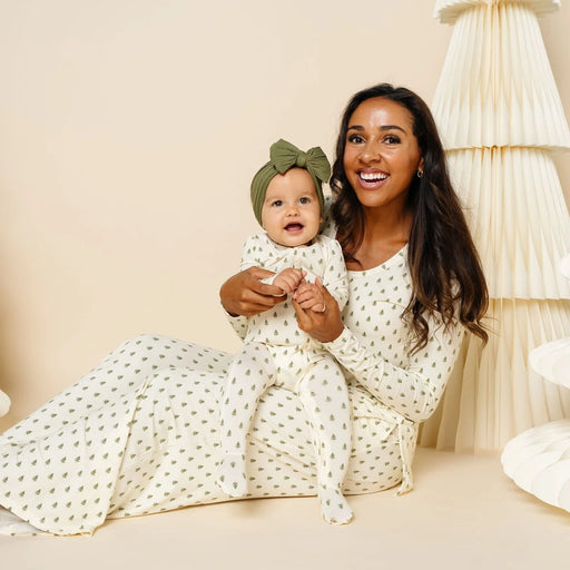 A woman and a baby sitting on a beige floor, both wearing matching cream-colored knit outfits with small green polka dots. The woman is holding the baby, who is wearing a matching green bow headband.