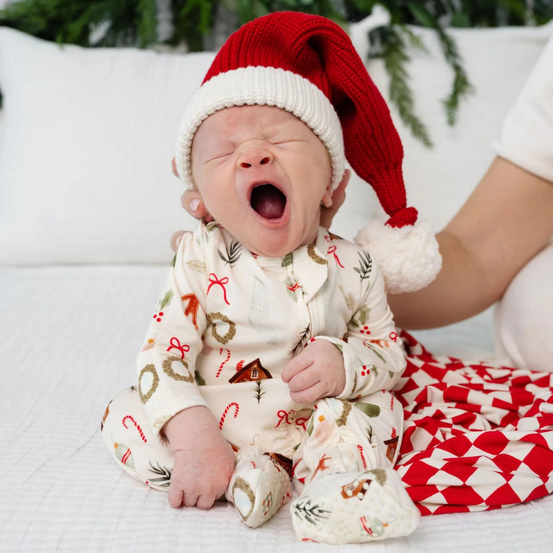 A newborn baby wearing a red Santa hat and a cream-colored Christmas-themed romper sits on a white quilted bed with a parent's hand gently touching the baby's shoulder.