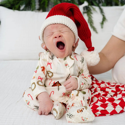 A newborn baby wearing a red Santa hat and a cream-colored Christmas-themed romper sits on a white quilted bed with a parent's hand gently touching the baby's shoulder.