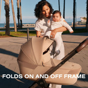 Mom holding baby beside Cybex Balios Fold Cot on stroller, folds on and off frame
