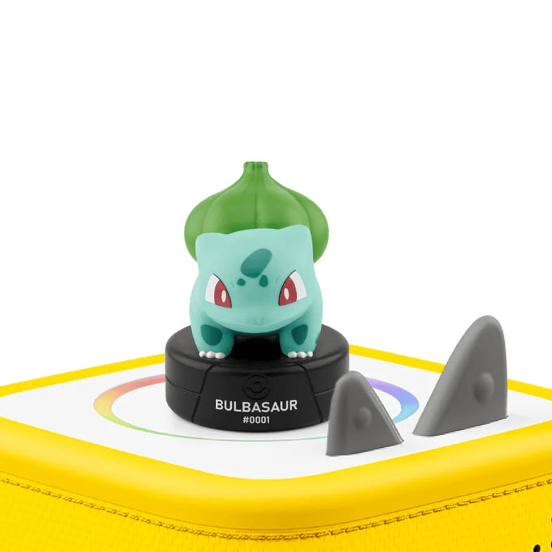 A Bulbasaur figurine from Pokemon, standing on a black circular base labeled 'BULBASAUR #2021', set on top of a yellow portable speaker with grey speaker cones visible.