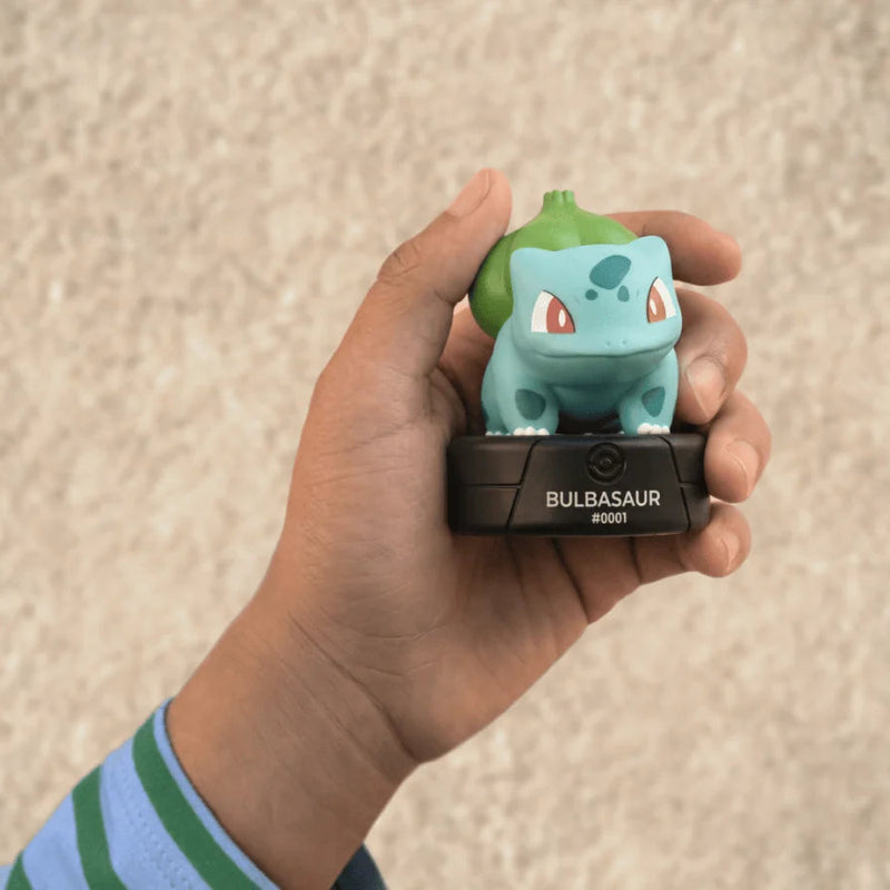 A hand holding a Bulbasaur amiibo figure on a black Nintendo Switch controller base, set against a textured beige wall.