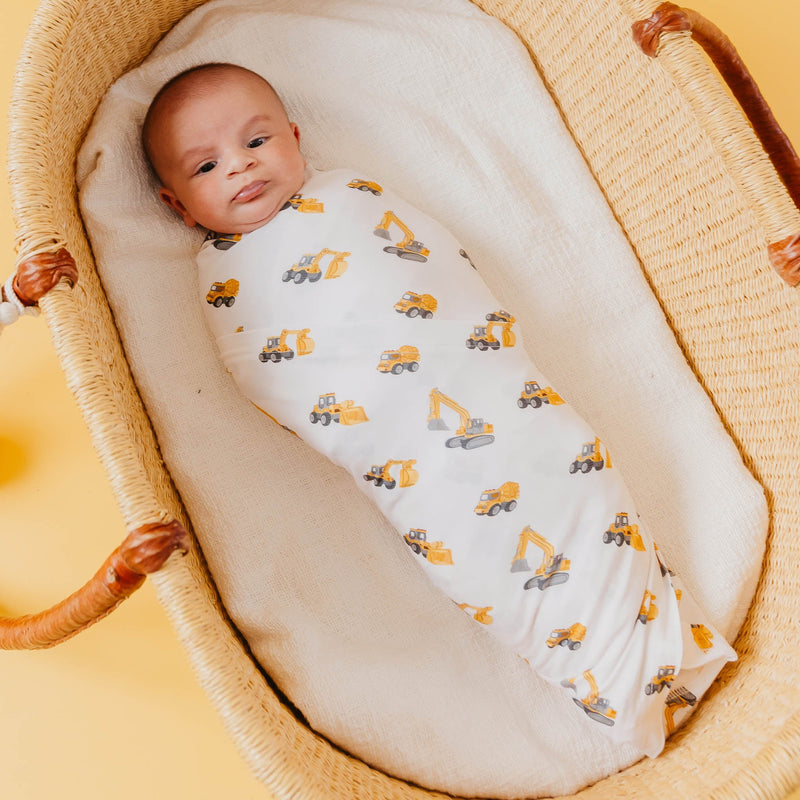 A baby swaddled in a white bamboo blanket with a yellow and gray construction vehicle print, lying in a woven rattan Moses basket.