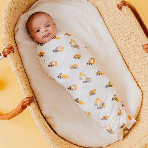 A baby swaddled in a white bamboo blanket with a yellow and gray construction vehicle print, lying in a woven rattan Moses basket.