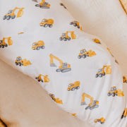 White baby bedding or a pillowcase printed with a repeating pattern of yellow construction vehicles, including excavators, dump trucks, and bulldozers, styled on a beige blanket.