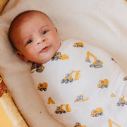 A baby swaddled in a white muslin blanket printed with yellow construction vehicles, lying in a woven basket.