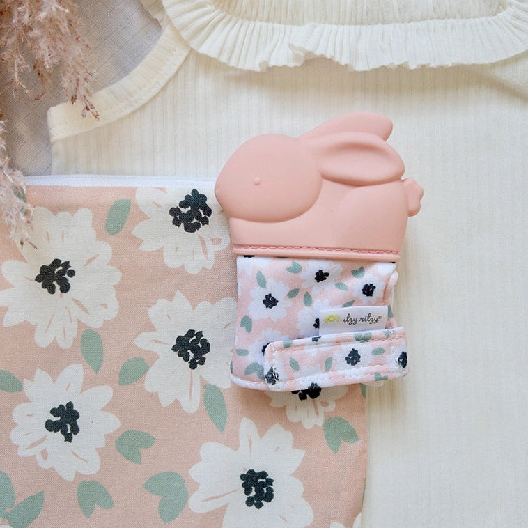 Silicone teether mitt displayed in a nursery setting on patterned fabric alongside baby accessories