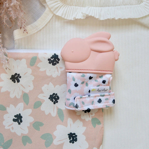 Silicone teether mitt displayed in a nursery setting on patterned fabric alongside baby accessories