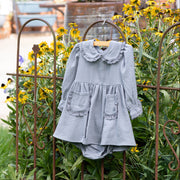 A light blue-gray long-sleeve baby dress with a ruffled Peter Pan collar and front patch pockets, hanging on a wooden hanger on a rustic metal garden fence in a garden setting with yellow flowers.