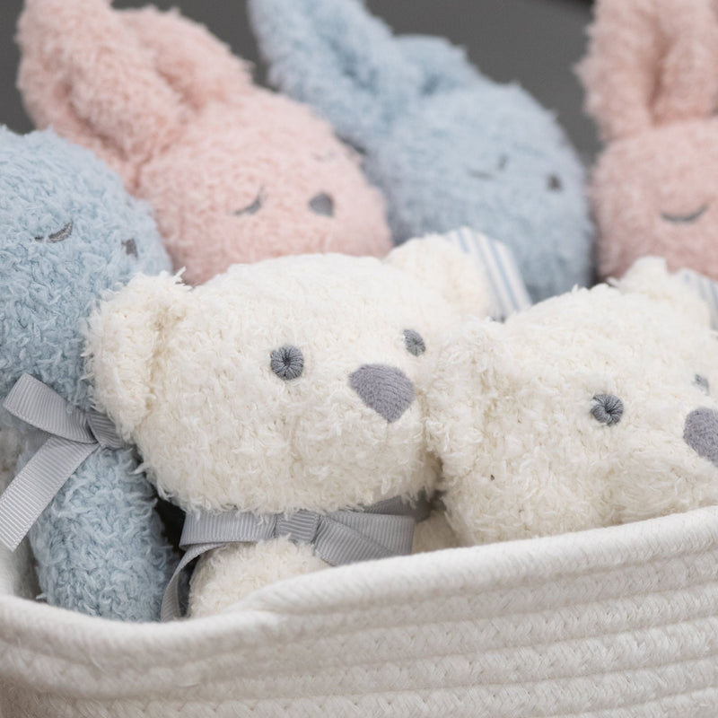 A basket of soft plush stuffed animals, including pastel blue and pink bunnies and cream-colored teddy bears, arranged in a white woven basket.
