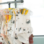 Baby bathing suits by milkbarn with coastal  prints on hangers against a neutral background
