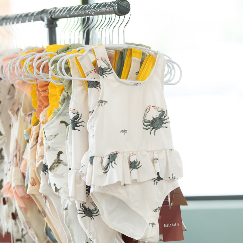 Baby bathing suits by milkbarn with coastal  prints on hangers against a neutral background