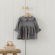 A gray baby girl's long-sleeve blouse with a ruffled Peter Pan collar and front patch pockets, hanging on a wooden hanger against a white paneled wall.