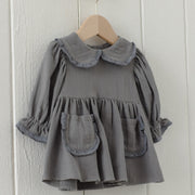 A toddler girl's gray linen dress with a ruffled Peter Pan collar, long sleeves with ruffled cuffs, and two front patch pockets, hanging on a wooden hanger against a white paneled wall.