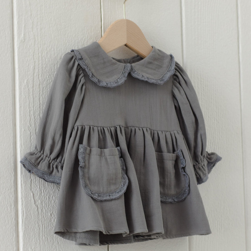 A toddler girl's gray linen dress with a ruffled Peter Pan collar, long sleeves with ruffled cuffs, and two front patch pockets, hanging on a wooden hanger against a white paneled wall.