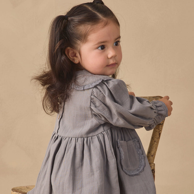 A young girl with dark hair in a ponytail, wearing a gray long-sleeve linen dress, posing on a rustic wooden chair against a warm beige background.