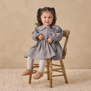 A young girl with dark hair in pigtails sits on a rustic wooden chair, wearing a dusty gray long-sleeve dress, white tights, and tan Mary Jane shoes against a neutral beige background.