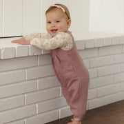 A baby standing on a dark wood floor and leaning on a white brick ledge, wearing a dusty rose overall jumpsuit over a cream floral long-sleeve shirt and a matching headband.