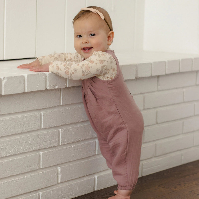 A baby standing on a dark wood floor and leaning on a white brick ledge, wearing a dusty rose overall jumpsuit over a cream floral long-sleeve shirt and a matching headband.