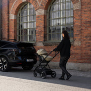 Parent pushing Thule Charm stroller with Thule Maple car seat past a brick building