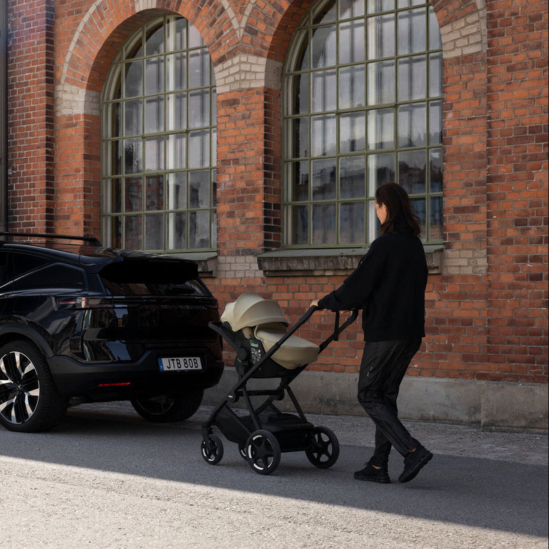 Parent pushing Thule Charm stroller with Thule Maple car seat past a brick building