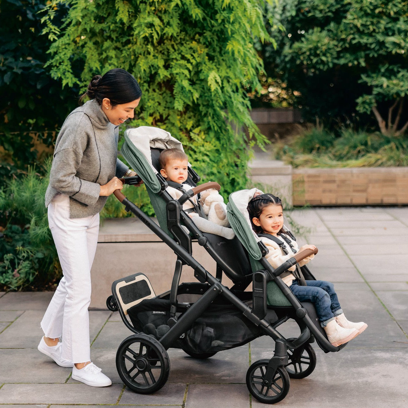 woman pushing baby and toddler uppababy vista v3 double stroller gwen green with ride on board piggyback