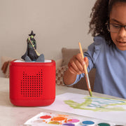 Child painting with a wicked witch on red tonies box red device and watercolors on a table.