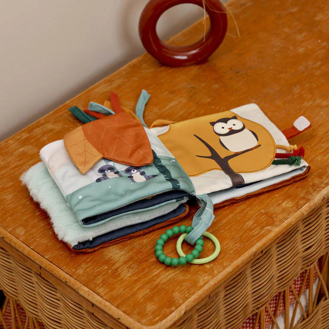 The soft book opened on a wooden dresser, showing forest-themed pages with fabric leaves, raccoons, and an owl illustration.