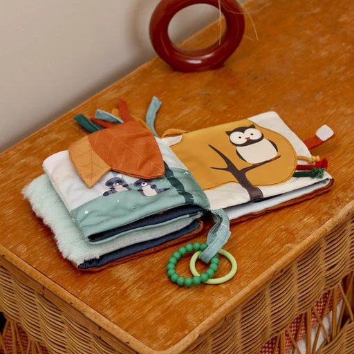 The soft book opened on a wooden dresser, showing forest-themed pages with fabric leaves, raccoons, and an owl illustration.