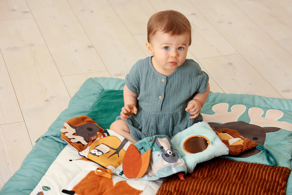 A baby sits on a forest-themed play mat while interacting with the activity book, surrounded by soft textures and woodland characters.
