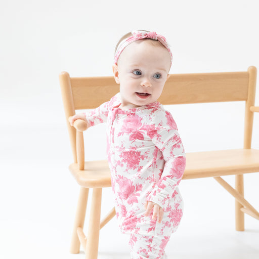 Baby wearing vintage pink roses zipper romper, standing beside a wooden chair