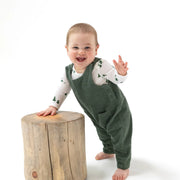 Toddler wearing dark ivy corduroy overalls over a printed long-sleeve tee, standing next to a wooden stump