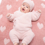A baby lying on a pink blanket with white heart patterns, wearing a fluffy light pink one-piece outfit and matching knit hat.