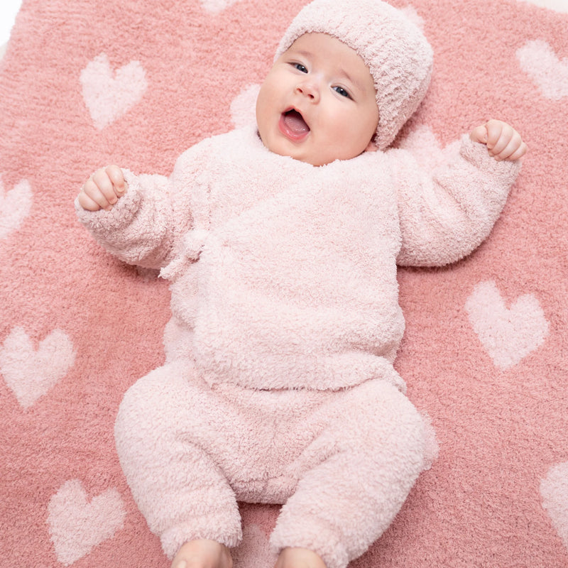 A baby lying on a pink blanket with white heart patterns, wearing a fluffy light pink one-piece outfit and matching knit hat.