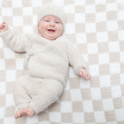 A smiling baby lying on a beige and white checkered blanket, wearing a fluffy cream-colored long-sleeve romper and matching beanie hat.