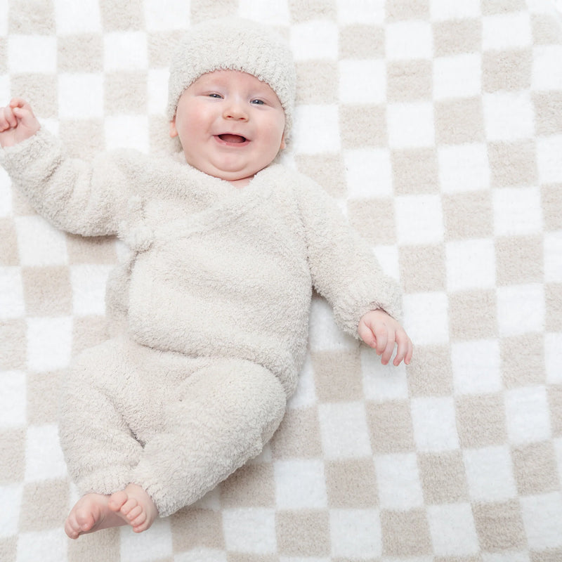 A smiling baby lying on a beige and white checkered blanket, wearing a fluffy cream-colored long-sleeve romper and matching beanie hat.
