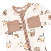 A close-up of a baby sleep sack or romper featuring a cute cartoon bubble tea and coffee cup pattern on an off-white background, with tan trim, a front zipper, and two patch pockets.