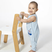 Child in a blue and white striped outfit with a sailboat design standing next to a wooden chair.