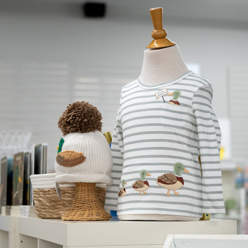 A children's long-sleeve striped shirt displayed on a mannequin torso, with duck illustrations on the front. A knitted duck hat sits beside it on a shelf in a softly lit indoor setting.